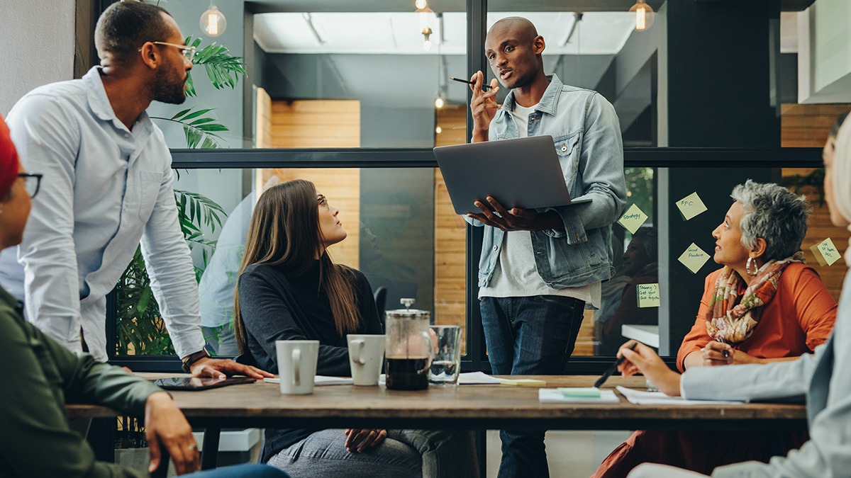 Five colleagues meet around a wooden table while one man stands and presents from a laptop to the group